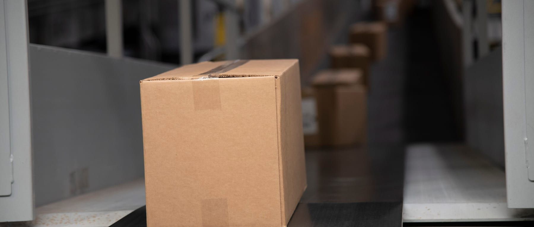 A cardboard box on a conveyor belt in a parcel depot, with more boxes receding out of focus behind it.