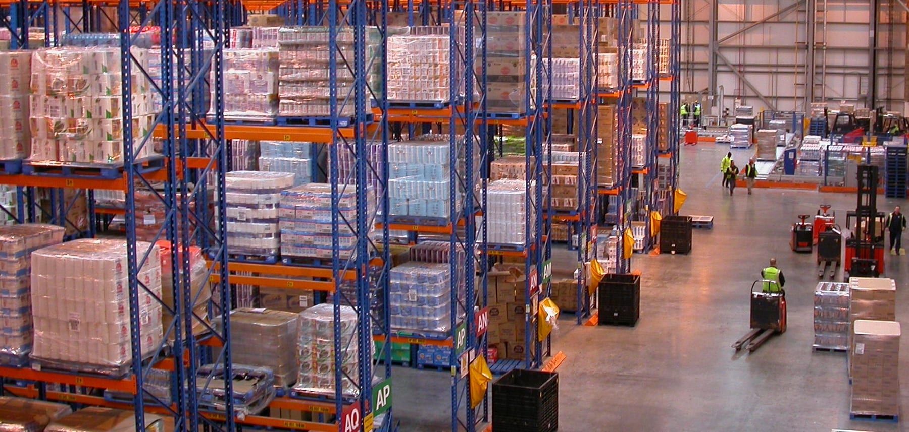 Pallet racking several storeys high in a distribution centre, with lettered aisle labels and workers moving pallet trucks along the floor.