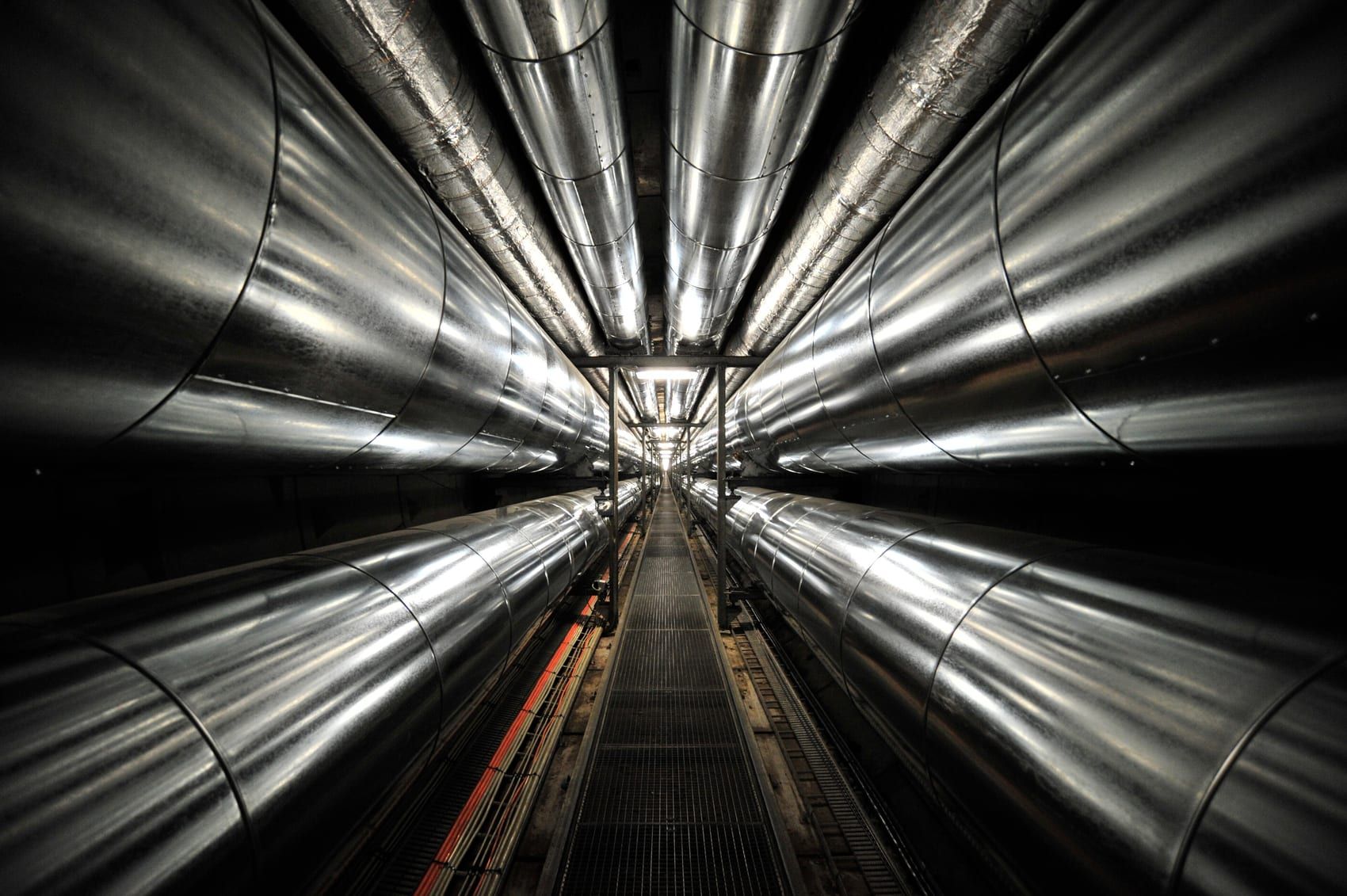 A narrow service tunnel lined on both sides with large insulated heating pipes, running away from the camera.