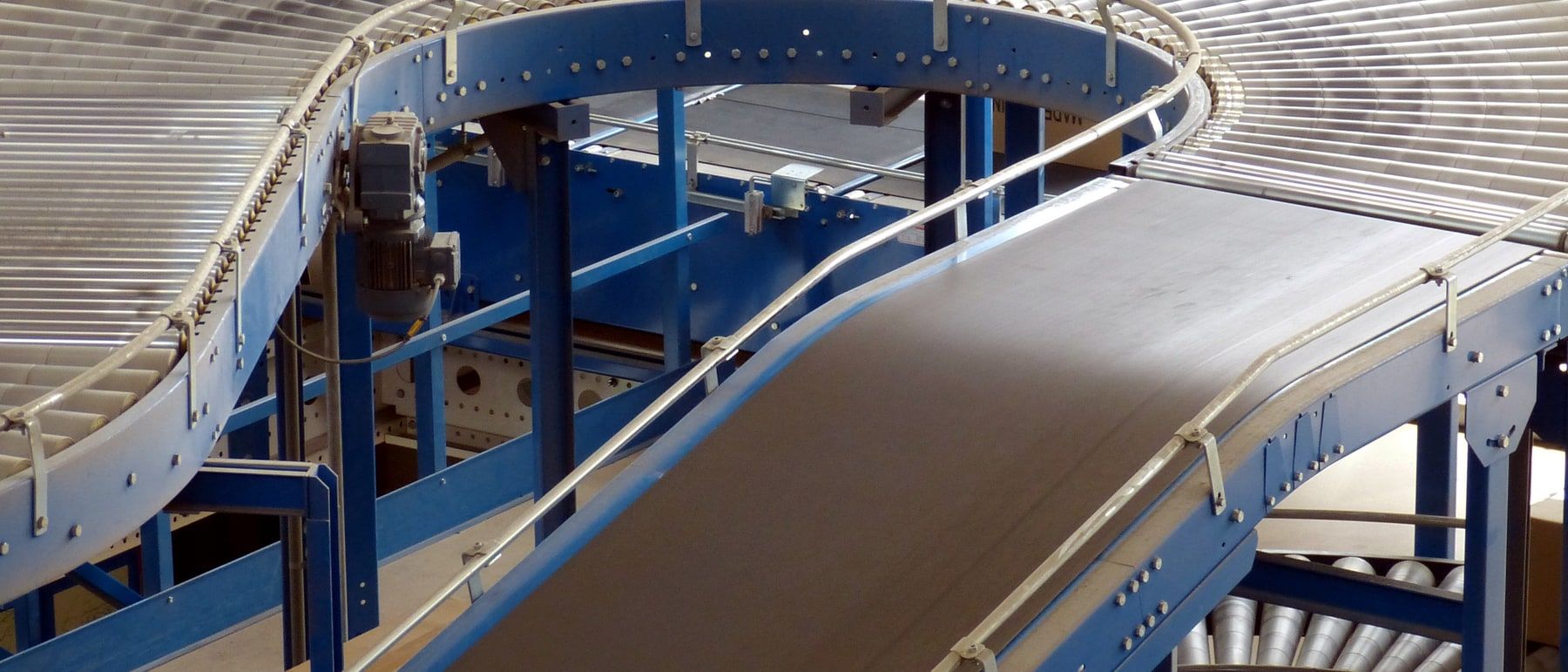 Powered roller conveyors curving through a sorting hall on blue steel legs, a belt section running into the bend and a second line crossing underneath.