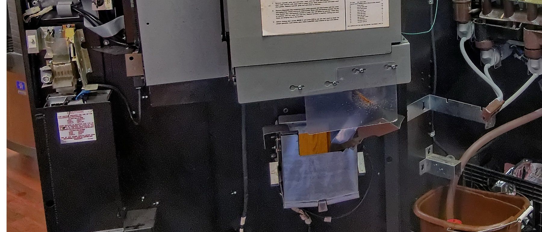 The open front of a coffee vending machine during a service visit: control boards and a coin unit on the door, tubing running down from the mixing heads, and a bucket on the floor catching what is flushed through.